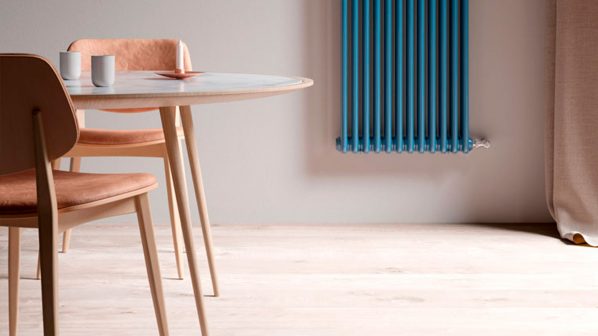 Un primo piano di un tavolo rotondo in legno chiaro con due sedie abbinate dalla seduta in pelle color cammello, posizionato su un pavimento in legno chiaro. Sul tavolo, un piatto con una decorazione rossa e due bicchieri bianchi. Sullo sfondo, si intravede un radiatore verticale a colonne di colore blu intenso, un modello di design Vasco, montato su una parete grigio chiaro. L'immagine suggerisce un ambiente domestico moderno e confortevole, con un'attenzione ai dettagli nel design degli arredi e degli elementi di riscaldamento.