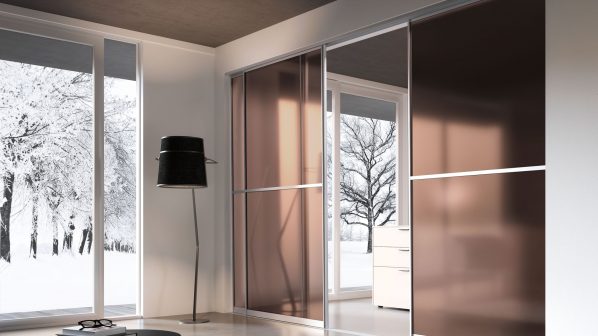 Una moderna cabina armadio o armadio con ante scorrevoli in vetro bronzo/rame trasparente del brand interno34, che riflettono il paesaggio esterno innevato attraverso ampie finestre. Le ante, con sottili profili in metallo, creano un ambiente luminoso e spazioso. In primo piano, un tavolino basso nero con un libro e occhiali è posizionato su un tappeto. Sullo sfondo, si intravede una cassettiera con una lampada da tavolo bianca. L'immagine mette in risalto il design elegante e la capacità di integrazione delle soluzioni di arredo di interno34 con l'ambiente esterno.