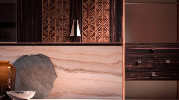 Un primo piano di una cucina o di una parete attrezzata del brand FMF, che mette in risalto l'uso di materiali pregiati e decorazioni sofisticate. La parete di fondo è rivestita in marmo rosato con venature scure e presenta un inserto roccioso naturale. Sopra, i pensili in legno scuro lucido con intarsi geometrici in metallo ramato. Sulla destra, una cassettiera in legno scuro con maniglie quadrate metalliche. Sulla sinistra, un vaso decorativo in ceramica e un altro oggetto cilindrico. L'illuminazione ambientale esalta la texture dei materiali. L'immagine enfatizza l'attenzione ai dettagli e il design di lusso tipici delle cucine FMF.