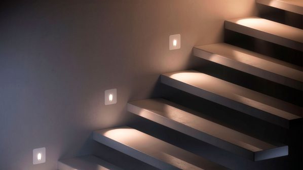 Una scala moderna con gradini chiari e una parete laterale che presenta tre lampade a incasso rettangolari, tipiche delle soluzioni di illuminazione per interni di Fabbian. Le luci emettono una luce bianca calda, creando un effetto di illuminazione d'accento sui gradini e lungo la parete, migliorando la visibilità e l'estetica della scala. L'immagine, con un'illuminazione soffusa generale, enfatizza il design minimale e la funzionalità delle luci segnapasso di Fabbian.
