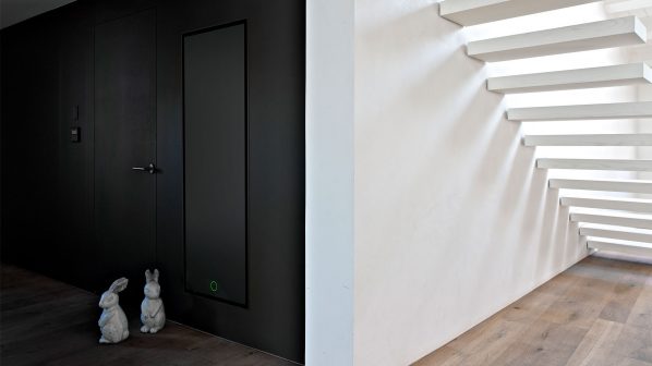 Un moderno radiatore di design verticale a incasso del brand Irsap, di colore nero opaco e a pannello liscio con un sottile profilo scuro, è integrato a filo in una parete nera in un ambiente contemporaneo. Il radiatore presenta un piccolo cerchio luminoso verde nella parte inferiore, che ne indica lo stato di funzionamento. A sinistra, una porta scura con maniglia nera si fonde con la parete, e due sculture decorative a forma di coniglietto bianco sono posizionate sul pavimento in legno scuro in primo piano. Sulla destra, una scala bianca dal design minimalista, con gradini che sembrano fluttuare, si innalza su una parete bianca, con il pavimento in parquet chiaro. L'immagine evidenzia l'integrazione discreta e il design sofisticato del radiatore Irsap in un ambiente moderno.