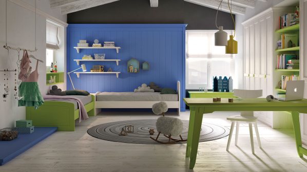 Una vivace e colorata camera per bambini del brand Scandola, pensata per ospitare due letti, con un'atmosfera moderna e funzionale. Sulla parete di fondo, un pannello blu intenso serve da testata per due letti singoli, uno verde lime a sinistra e uno bianco al centro, entrambi con cuscini grigi e bianchi. Sopra i letti, mensole a muro bianche con oggetti decorativi e libri. Sulla destra, una libreria bianca a scaffali aperti, in parte riempita con libri e una piccola lampada verde, si trova accanto a una scrivania verde lime con un laptop aperto e una sedia bianca. Il pavimento è in legno chiaro. In primo piano, un tappeto rotondo grigio con disegni concentrici e un dondolo a forma di pecora aggiungono un tocco giocoso. Sulla sinistra, un appendiabiti a parete con indumenti e un materassino blu completano l'ambiente. La luce naturale proviene da ampie finestre, e il soffitto spiovente con travi a vista aggiunge carattere alla stanza.
