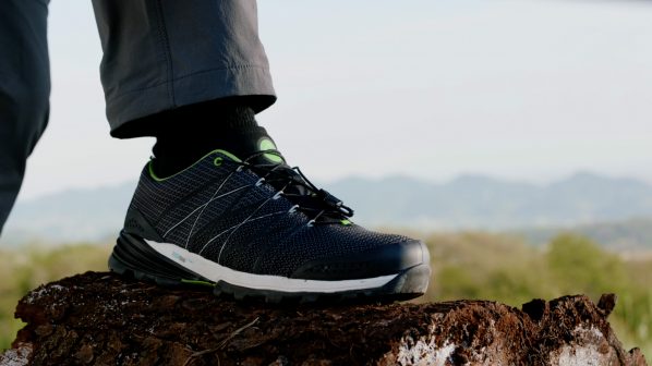 Un primo piano della parte inferiore di una gamba di una persona che indossa pantaloni grigi scuri e una scarpa sportiva da trekking. La scarpa è di colore nero con dettagli verdi brillanti e lacci bianchi che creano un motivo intricato. È appoggiata su un ceppo d'albero scuro e irregolare. Lo sfondo è sfocato e mostra un paesaggio montano con cielo chiaro e vegetazione. L'immagine è scattata all'aperto, probabilmente in un ambiente naturale.