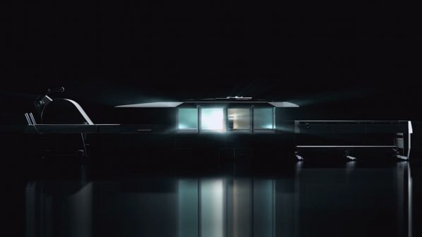 Una macchina industriale LXN REVOLUTION di Dallan è parzialmente visibile in un ambiente buio, con le sue luci interne e le finestre che emettono un bagliore bluastro e bianco. La macchina ha una forma allungata e complessa, con sezioni che suggeriscono una linea di produzione. Dettagli sul lato sinistro sembrano essere componenti meccanici o un braccio robotico, mentre la sezione centrale è illuminata come un'area di lavoro o di osservazione. La superficie scura sottostante riflette le luci della macchina, creando un effetto futuristico e misterioso. L'immagine suggerisce che la macchina è potente e tecnologicamente avanzata.