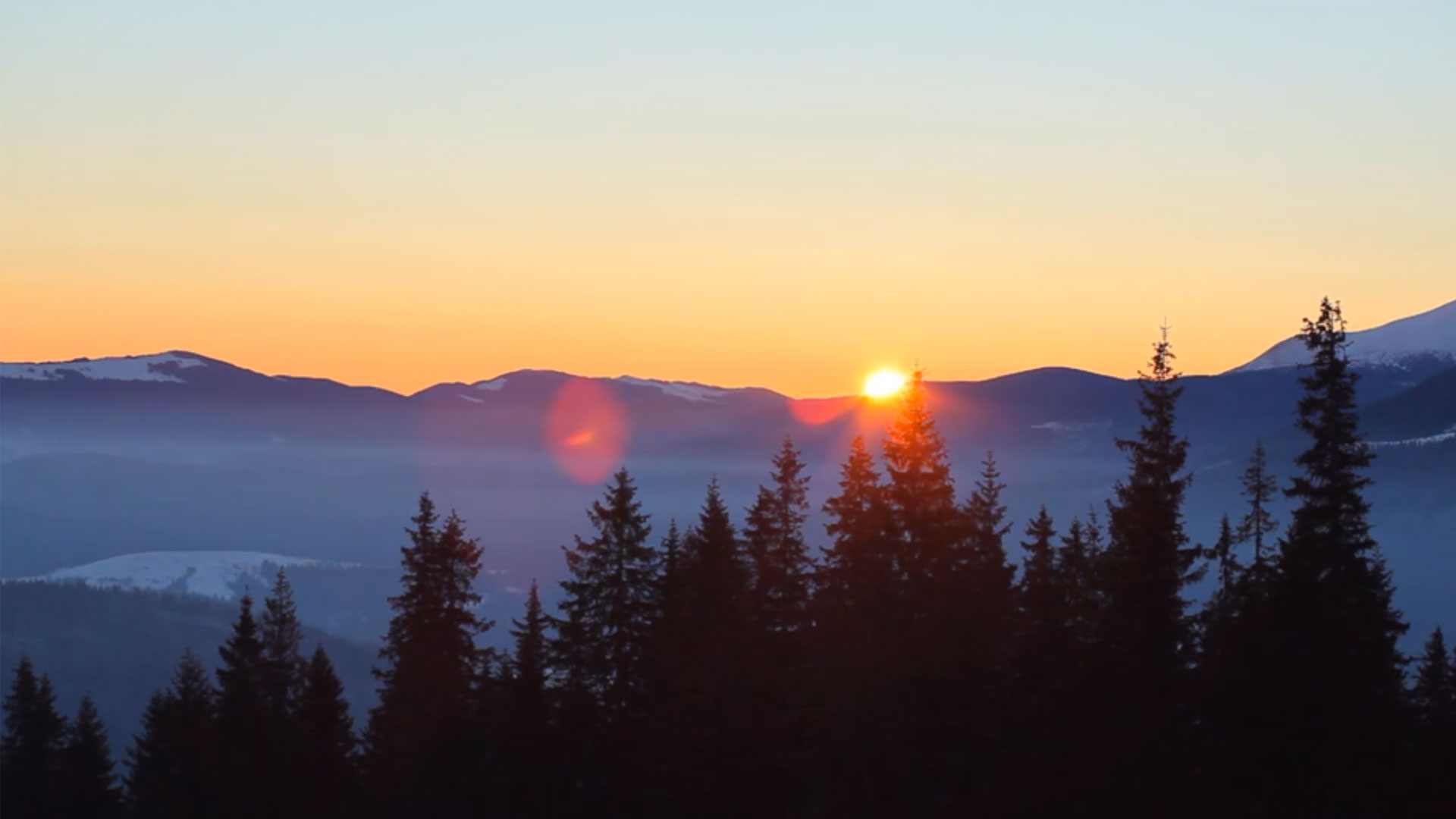 Un suggestivo tramonto sulle montagne, con il sole arancione brillante che si appoggia appena dietro una catena di montagne scure e sfumate in lontananza. Le cime più alte a destra mostrano tracce di neve. In primo piano, le sagome scure e appuntite di numerosi abeti si stagliano contro il cielo che sfuma dall'arancione all'azzurro chiaro verso l'alto. Una leggera foschia bluastra avvolge le valli tra le montagne.
