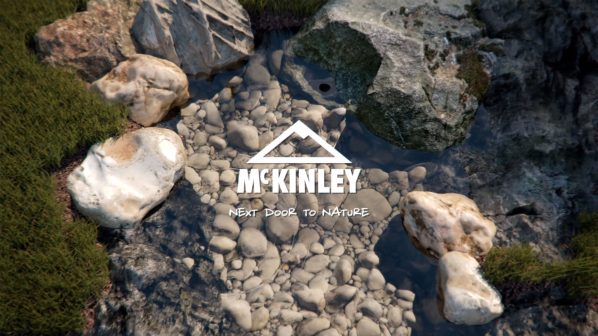 Una vista dall'alto di un ruscello poco profondo che scorre su un letto di ciottoli e rocce levigate. L'acqua è limpida e riflette leggermente il cielo. Ai bordi del ruscello, cresce dell'erba verde e muschio. Al centro dell'immagine, sovrapposto all'acqua e alle rocce, è posizionato il logo bianco di "MCKINLEY", con un simbolo stilizzato di una montagna sopra il nome. Sotto il logo, la frase "NEXT DOOR TO NATURE" (Accanto alla Natura) è visibile, rafforzando il tema naturale dell'immagine.