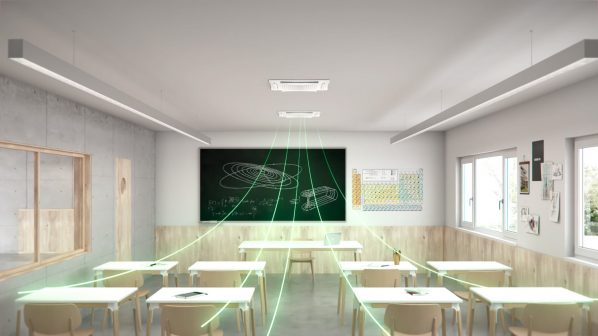 Un'aula di una classe moderna, illuminata da luci a LED rettangolari incassate nel soffitto bianco. Due unità di purificazione dell'aria Sabiana SkySafe MD600 sono visibili incassate nel soffitto, con le loro griglie quadrate. Dalle unità, raggi di luce verde simboleggiano il flusso d'aria purificata, che si estende verso i banchi chiari e le sedie di legno disposti ordinatamente nella stanza. Sullo sfondo, una lavagna nera mostra diagrammi e formule con scritte verdi, mentre sulla parete destra è appeso un grande poster della tavola periodica. La parete di sinistra ha un aspetto grezzo in cemento, mentre quella di destra è rivestita in legno chiaro nella parte inferiore.