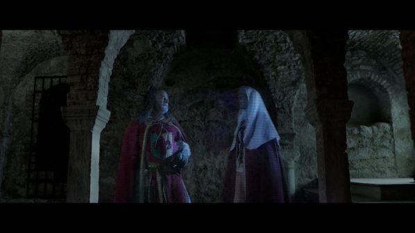 Un'immagine buia e suggestiva che raffigura un ambiente sotterraneo o una cripta, illuminata da una luce fioca che mette in risalto le texture grezze delle pareti e degli archi. In primo piano, due figure translucide e spettrali sono delineate da una leggera aura bluastra e da sottili linee che ne suggeriscono la forma. La figura a sinistra, in abiti che ricordano un'armatura rossa, sembra reggere qualcosa. La figura a destra, con un copricapo bianco e un vestito scuro, è rivolta verso l'altra. L'atmosfera è misteriosa e inquietante, con dettagli architettonici in pietra, come archi e colonne, che si perdono nell'ombra profonda.