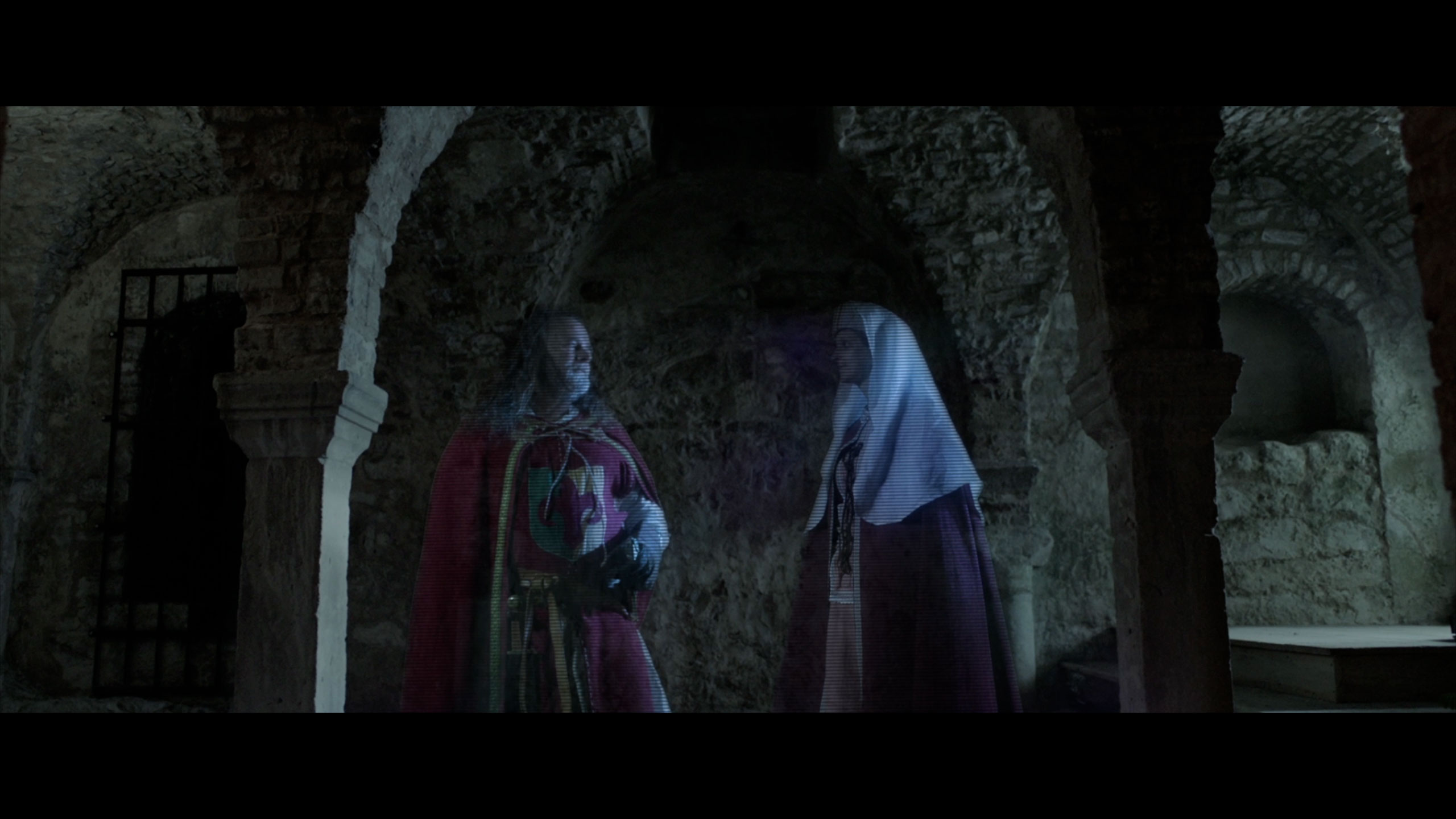 Un'immagine buia e suggestiva che raffigura un ambiente sotterraneo o una cripta, illuminata da una luce fioca che mette in risalto le texture grezze delle pareti e degli archi. In primo piano, due figure translucide e spettrali sono delineate da una leggera aura bluastra e da sottili linee che ne suggeriscono la forma. La figura a sinistra, in abiti che ricordano un'armatura rossa, sembra reggere qualcosa. La figura a destra, con un copricapo bianco e un vestito scuro, è rivolta verso l'altra. L'atmosfera è misteriosa e inquietante, con dettagli architettonici in pietra, come archi e colonne, che si perdono nell'ombra profonda.