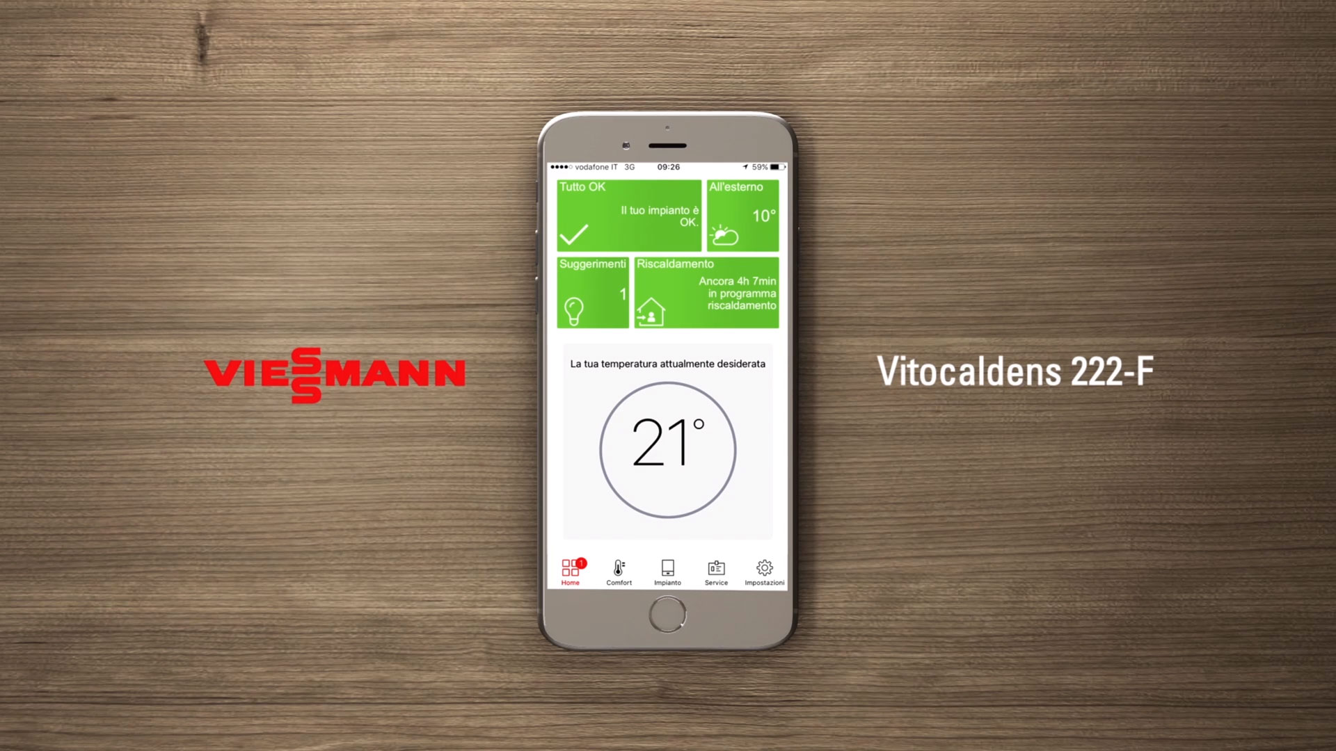 Uno smartphone bianco è posizionato verticalmente su una superficie di legno scuro e lucido, mostrando lo schermo di un'applicazione per il controllo della temperatura e del riscaldamento. L'app visualizza "Tutto OK" e "Il tuo impianto è OK" in verde in alto, e una temperatura esterna di "10°". Al centro, un grande indicatore circolare mostra la temperatura desiderata di "21°". In basso, il logo "VIESSMANN" è visibile sulla sinistra e "Vitocalcdens 222-F" sulla destra. L'interfaccia dell'app include anche icone per "Home", "Contact", "Service" e "Impostazioni".