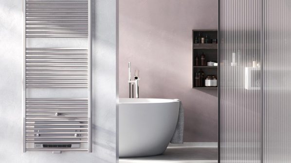 Un moderno scaldasalviette a parete di colore argento lucido, con barre orizzontali e piatte, è installato su una parete grigia chiara. Sulla parte inferiore dello scaldasalviette, un termostato digitale è visibile. A destra, una vasca da bagno bianca lucida con un rubinetto cromato. Accanto alla vasca, una porta scorrevole in vetro trasparente con un motivo a strisce verticali. Sullo sfondo, una parete rosa chiaro con una mensola nera a cubi contenente oggetti decorativi. Il pavimento è in cemento lucido.