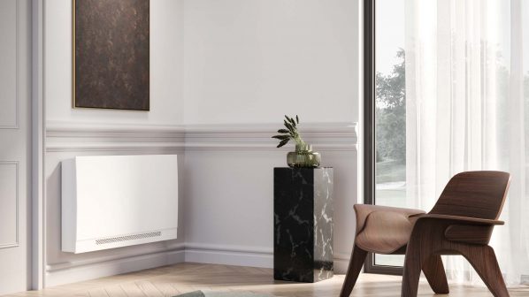 Un moderno termoconvettore bianco è montato su una parete chiara con modanature decorative, in un angolo di una stanza. A sinistra, si intravede una porta bianca semi-aperta. Sopra il termoconvettore, un quadro rettangolare verticale con una superficie marrone scuro. A destra, lunghe tende grigie e un piedistallo alto di marmo nero con una pianta verde. In primo piano a destra, una sedia di design in legno scuro con schienale e seduta curvi. Il pavimento è in parquet di legno chiaro. Si intravede un tappeto geometrico con sfumature di grigio e verde in primo piano a sinistra.