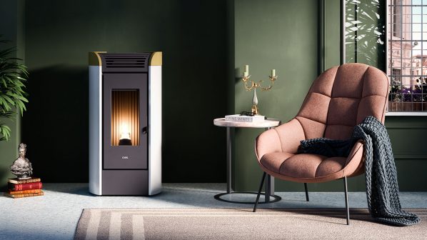 Una stufa a pellet di colore nero e grigio chiaro con dettagli color legno sulla parte superiore, posizionata in un moderno soggiorno con pareti verde scuro. La stufa ha una finestra verticale che mostra una fiamma accesa e il logo "CADEL" in basso. A sinistra, una grande pianta verde in un vaso bianco e una scultura su una pila di libri. A destra, una poltrona imbottita rosa chiaro con un plaid blu scuro e un tavolino rotondo bianco con un candelabro dorato. Una finestra con persiane in legno è visibile sullo sfondo a destra.