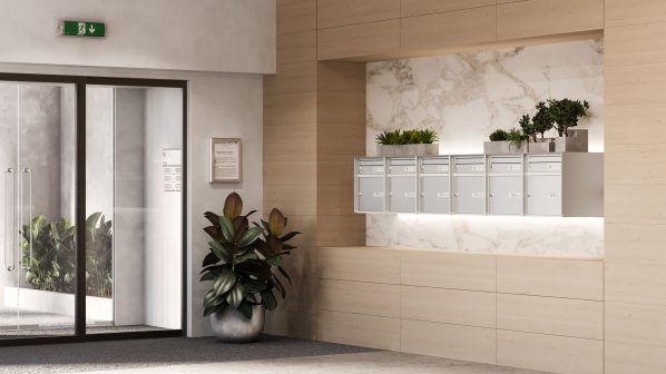 L'ingresso di un moderno edificio con una pavimentazione chiara e pareti con pannelli in legno chiaro. A destra, in una nicchia rivestita in marmo bianco e grigio, si trovano diverse cassette postali orizzontali di colore argento. Sopra di esse, piante verdi decorano la mensola. In primo piano a sinistra, una grande pianta verde in un vaso grigio lucido. Sulla sinistra, doppie porte a vetro con telai neri conducono all'interno dell'edificio.