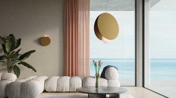 Un elegante salotto moderno con un divano modulare bianco imbottito e un tavolino da caffè rotondo in marmo grigio. Sul tavolino, un vaso con fiori rosa. Una grande lampada a sospensione di colore dorato con un globo luminoso bianco pende dal soffitto. Le pareti sono chiare e una tenda rosa voluminosa è drappeggiata a sinistra di un'ampia finestra che si affaccia sul mare.