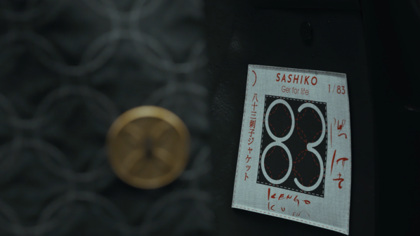 Un primo piano di un'etichetta bianca rettangolare con un bordo cucito e un motivo nero che assomiglia al numero "83" in un cerchio. Sopra il motivo, la parola "SASHIKO" e "Get for life" con "1/83" nell'angolo in alto a destra. Testo in giapponese è presente sul lato sinistro dell'etichetta. L'etichetta è posta su uno sfondo scuro con un motivo sfocato di cerchi e una forma circolare dorata sfocata in alto a sinistra.