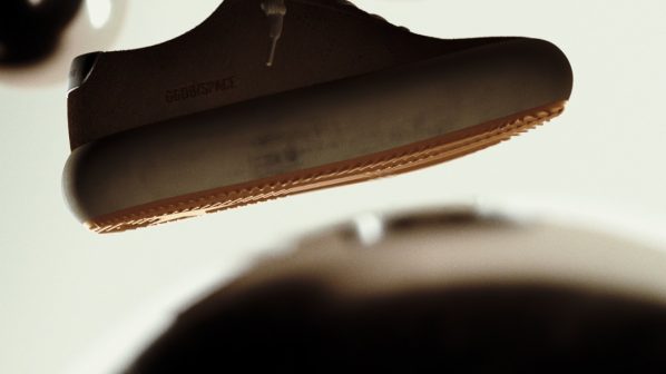 Una scarpa sportiva casual scura, probabilmente marrone o nera, con suola spessa e lacci chiari, fluttua al centro di un ambiente astratto. Grandi sfere scure e sfocate si trovano in primo piano e sullo sfondo, creando un'atmosfera minimalista.