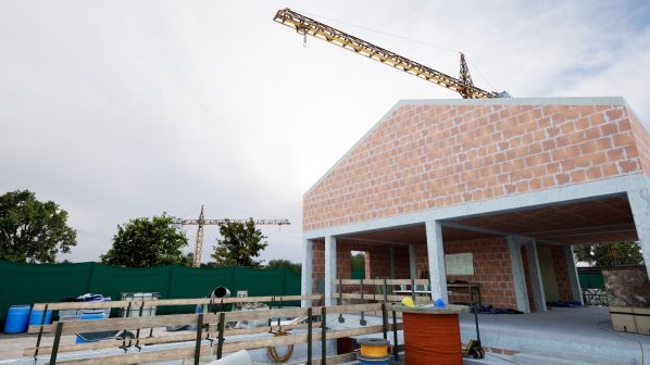 Un edificio in costruzione con pareti in mattoni a vista e struttura in cemento armato, sormontato da una grande gru edile gialla. In primo piano si vedono barili blu, recinzioni in legno e bobine di cavi. Sullo sfondo, reti di sicurezza verdi e alberi sotto un cielo nuvoloso.